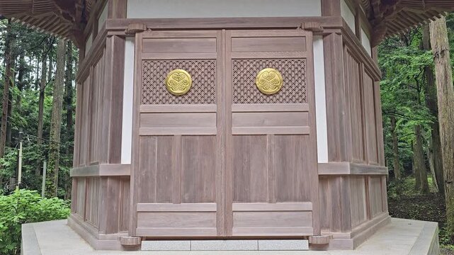 Traditional Japanese wooden shrine building with carved entrance doors and golden family crest surrounded by forest. Static architectural footage of historic temple in natural woodland setting