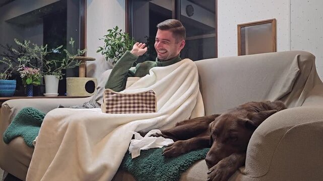 Adult man wrapped in blanket laughing after dog passes gas and fanning away unpleasant smell while sitting on sofa at home with chocolate Labrador. Funny pet behavior, humor, friendship