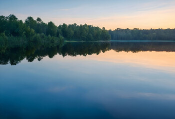 Obraz premium Calm lake with forest reflection at sunrise, tranquil nature landscape with clear sky and still water, the clear sky transitions from blue to orange and yellow hues