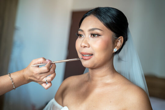 Close up of an elegant Asian bride with braces getting her lipstick applied by a makeup artist during wedding day preparation. Beautiful bridal makeup detail.