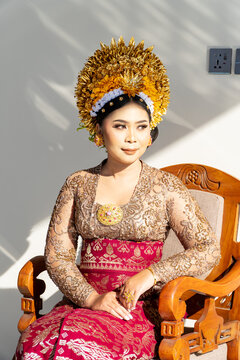 Beautiful portrait of a Balinese bride in traditional payas agung attire, featuring a golden crown and lace kebaya, sitting in natural sunlight.