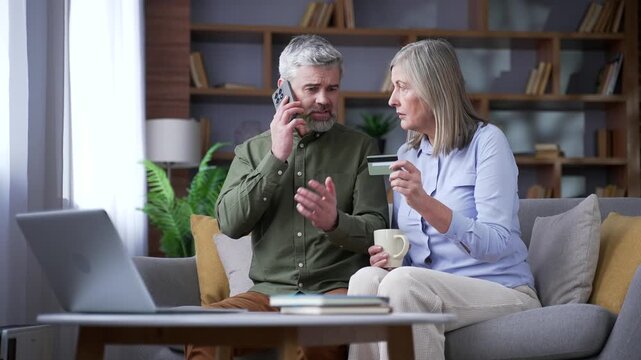 Angry senior man talking on a smartphone while sitting on a sofa beside his wife in the living room at home. Retired couple dealing with a credit card problem as he calls the bank for assistance.