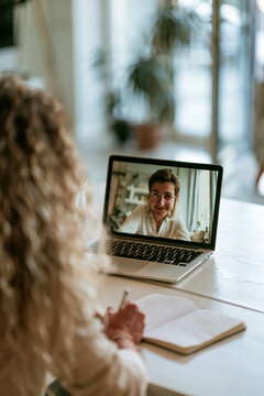 A candidate is sitting in a clean, minimalist home office, looking at a laptop screen where a recruiter's face is visible in a video call.