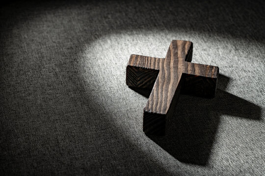 Wooden christian cross with dramatic light and shadow. Closeup of textured wooden cross lying on gray fabric under dramatic spotlight beam creating deep shadow for religious and spiritual concepts
