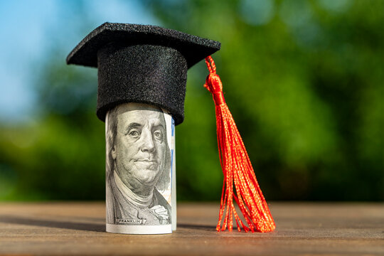 Graduation cap on US dollar bill roll. Miniature academic cap on rolled hundred dollar banknote for tuition fees student loan investment in education and high cost university concepts