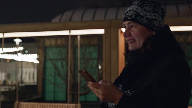 Smiling woman in dark coat, winter hat holds smartphone while standing outside illuminated storefront at evening, checking messages or browsing.