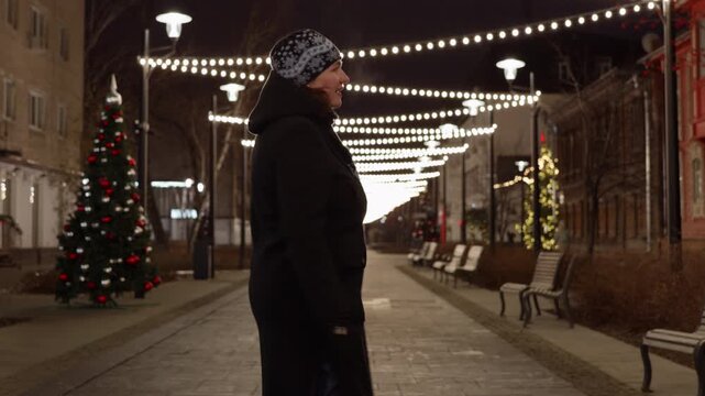 Woman in black coat, knit hat walks through decorated urban street at night, smiling at illuminated Christmas lights strung overhead, evergreen trees with ornaments lining sidewalk.