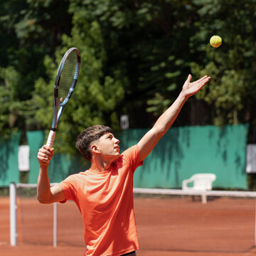 Teenage tennis player tossing the ball before serving on outdoor clay court, focus, preparation and serving technique concept. Young male athlete with tennis racket and ball performing service