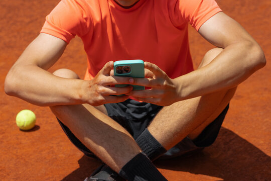 Teenage tennis player using smartphone while resting on outdoor clay court, digital communication, recovery break and modern sports lifestyle