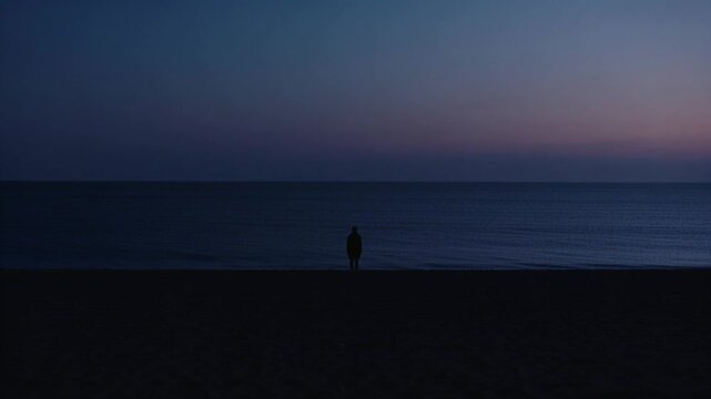 Standing man in dark coat watching horizon beachside as light shifts seeing sunrise with calm ocean