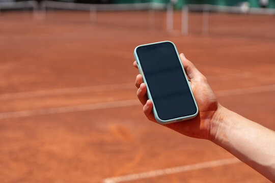 Smartphone with blank screen held over clay tennis court, ideal mockup for sports apps, tournament information, coaching services and mobile advertising