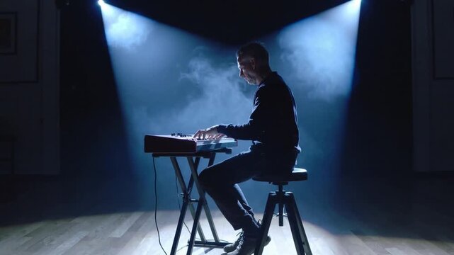 Pulling camera back, adult male musician wearing dark sweater playing synth on studio stage for set