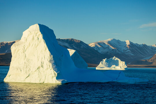 Two large icebergs drift in blue Arctic waters beneath snow-covered mountains near Ittoqqortoormiit, Greenland