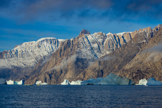 Bright blue icebergs outline rock faces of warming islands mountain range under clear skies in Greenland