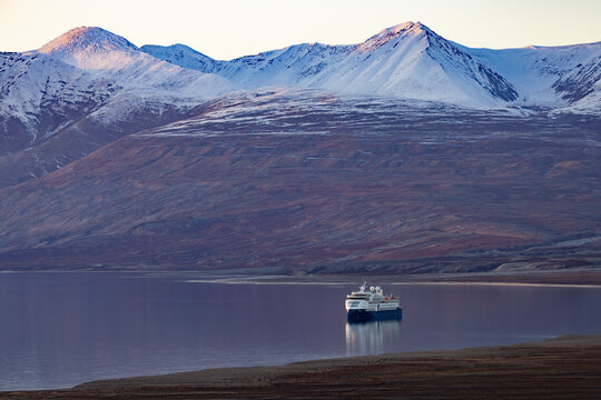 Expedition vessel anchors in golden waters under mountains at dusk in Blomsterbugten, Greenland