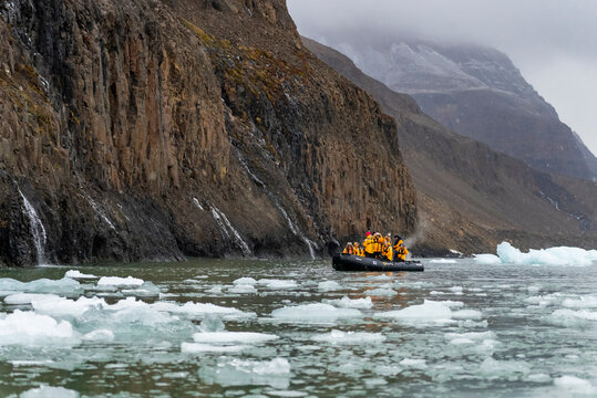 Zodiac boat explores icy waters with tourists in yellow parkas navigating beneath cliffs in Borgvig, Greenland