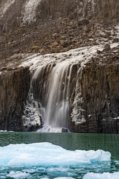 Tall waterfall pours over dark basalt cliffs into icy green water with blue ice pieces floating near the base in Greenland