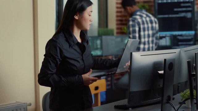 Asian software developer approaches an agency workstation with a coded laptop interface, connecting portable computing, app development and office workflow