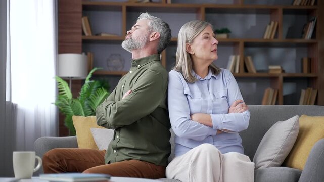 Senior married couple sitting on a sofa in the living room at home with crossed arms after an argument. Retired husband and wife avoiding each other as relationship conflict creates emotional distance