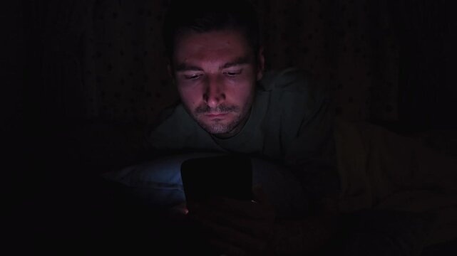 Man using smartphone in bed at night in dark bedroom. Close up footage illuminated by phone screen, showing late night mobile use, screen time, sleeplessness and digital lifestyle