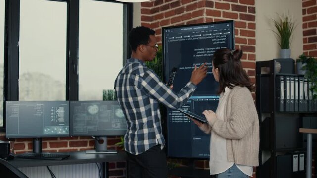 Software developer analyzes programming issues on a large office display with a digital tablet, reviewing bugs, inside a technical startup workspace.
