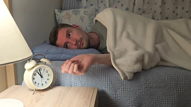 Young man lying in bed sets alarm clock before sleep for early morning work, switches off bedside lamp and falls asleep. Evening bedtime routine, preparing for workday, healthy sleep schedule