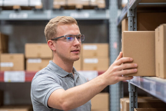 A young blond man wearing glasses reaches to pick up a cardboard box from a metal storage shelf in a busy warehouse.