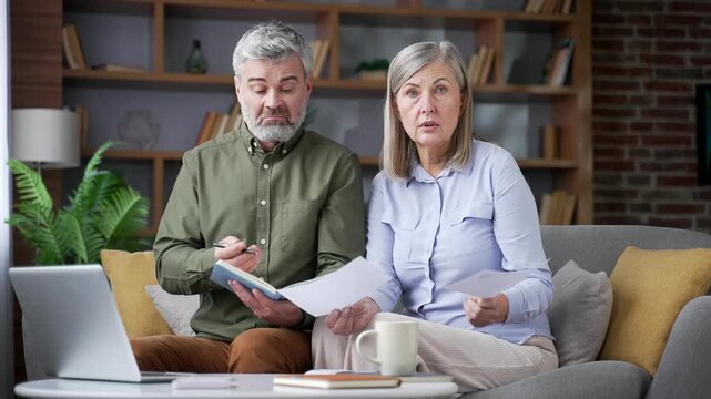 Confused senior married couple holding utility bills and a notebook while managing household finances in a home office. Retired husband and wife then looking at the camera with worried expressions.