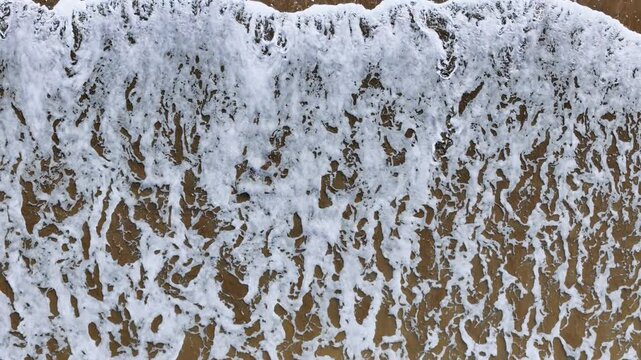 Top down aerial view of foamy waves washing onto sandy beach. Natural wave texture and coastal background