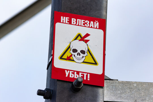 Close-up view of a red and yellow danger sign on a metal utility pole, featuring a skull icon and a Russian warning text about high voltage electric shock hazards, Russia