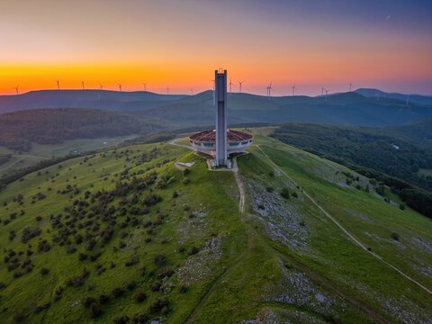 Drone view of Buzludzha monument atop a mountain during sunset with wind turbines in the distance in Bulgaria