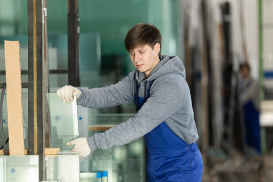 In glass workshop, young man glazer works with glass sheets and related supporting parts, preparing PVC products for interior finishing work.