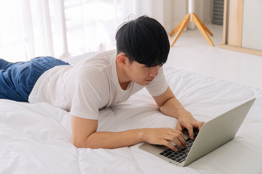 Focused young Asian man lying comfortably on a white bed, typing on a laptop. Dedicated freelancer or student working from home in a bright, cozy bedroom. Remote digital lifestyle.