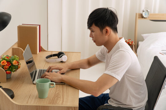Focused young Asian man working on laptop at wooden desk in bright bedroom. Dedicated freelancer or student studying online at home with coffee and headphones.