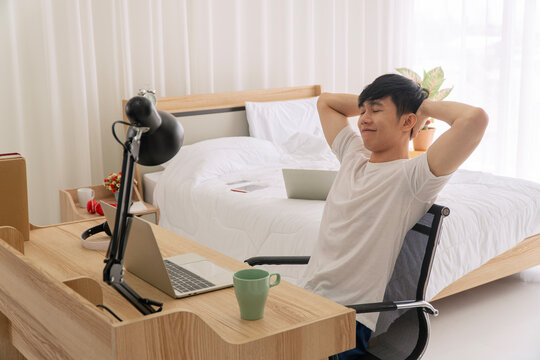 Smiling young Asian man relaxing with hands behind his head after finishing work on a laptop at a wooden desk. Happy freelancer taking a break in a bright, comfortable bedroom home office.