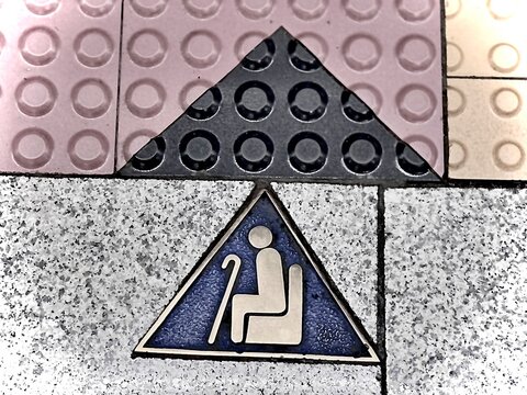 Close-up of tactile paving blocks with a triangular tile icon showing an elderly person with a cane, indicating priority or caution area for disabled and elderly pedestrians.