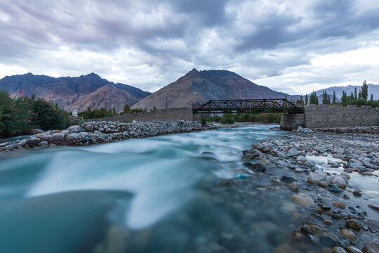 Bridge crosses turquoise glacial river in Nubra Valley with Himalayan peaks in background during overcast sky