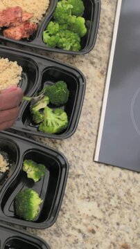Vertical video: Spooning hand filling trays with broccoli by stove for meal prep with rice, chicken