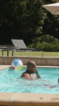 Vertical video: Rising, splashing man in trunks swimming in home pool, having fun with beach ball