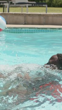 Vertical video: Playing dad-son at pool in red trunks, son nudging, dropping and swimming with ball