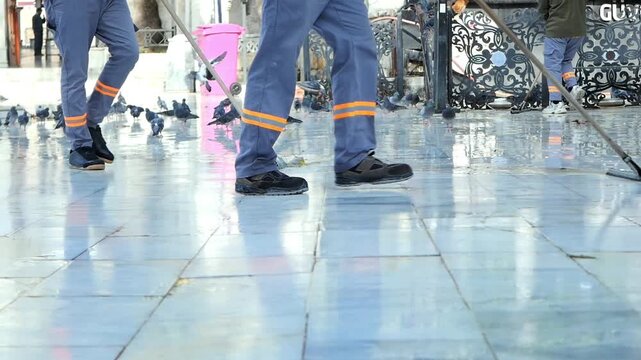 Cleaners sweeping wet tiled floor with long handled squeegees while pigeons walk around in public space low angle