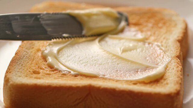 Spreading person's hand dragging serrated knife across butter blob on toast on plate for breakfast