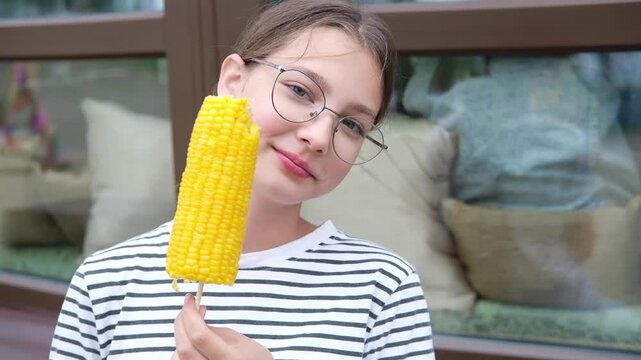 Young Caucasian girl with glasses smiles while holding a corn on a stick, showcasing enjoyment in a cozy outdoor setting with cushions in the background