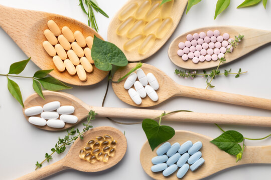 Wooden spoons holding various health supplements, soft gels, capsules, tablets, on white background.  Fresh green leaves, vines, and herb accents.