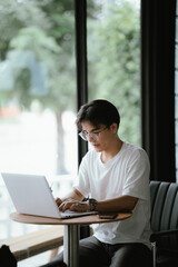 Fototapeta premium Young professional talking on smartphone while working on a laptop in a modern café, holding a coffee cup with natural window light.