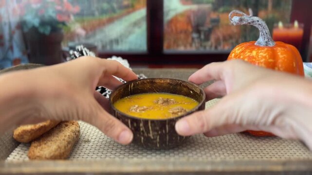 Manos sosteniendo un cuenco de sopa de calabaza junto a la ventana