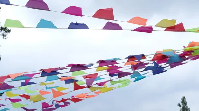 Colorful triangular flags in vibrant hues sway gently in the breeze against a cloudy sky, creating a festive atmosphere in an outdoor setting with trees in the background