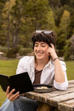 Woman sits at a wooden picnic table in a park. She wears a white shirt and jeans, with sunglasses on her head. She smiles while holding a book. Plate with food and a drink sits beside her