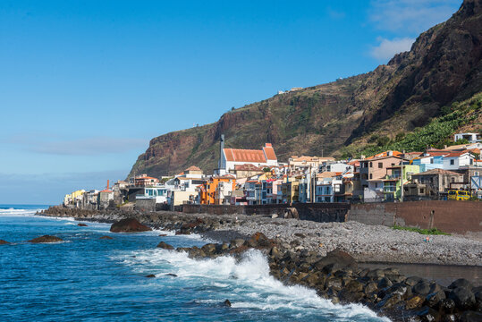 Colorful buildings line the coast of Paul do Mar in Madeira, with waves crashing against the rocks and a blue sky above