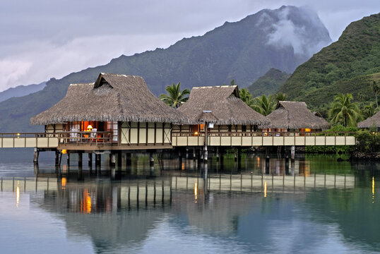 Bungalows on stilts in Moorea reflecting in water at dusk, South Pacific Ocean